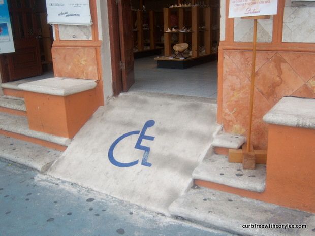 Eine kurze, steile Betonrampe mit einem aufgemalten Rollstuhlsymbol führt vom Bürgersteig hinauf zu einer offenen Tür eines Ladens